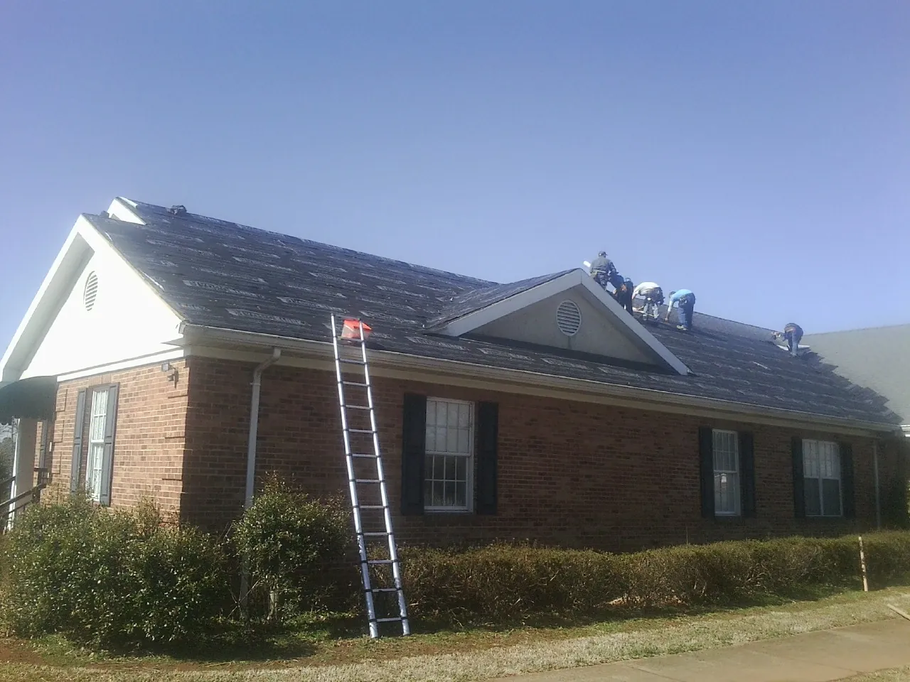 Crew tearing shingles off a brick ranch roof with a ladder set — Metal Roof Repair work by our crew in Springport