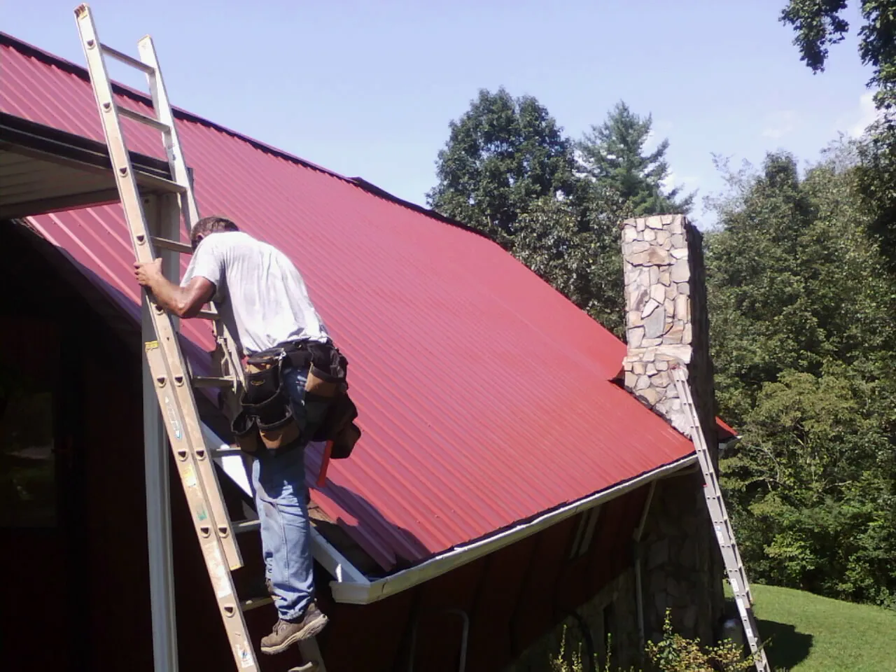 Roofer on a ladder beside a red metal roof and stone chimney — Soffit & Fascia Repair by Roofing Brothers in Springport